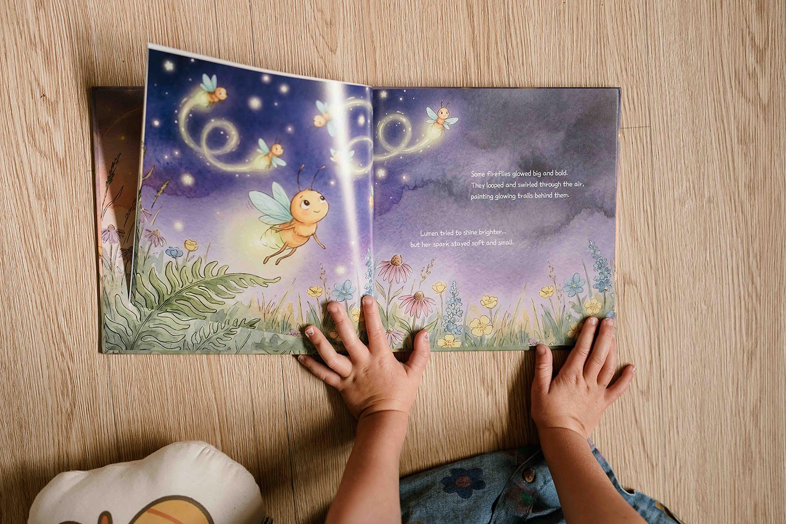 Overhead photograph of a child reading an open copy of The Smallest Spark, showing a purple illustrated spread filled with glowing fireflies